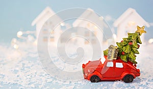 Christmas decor - red retro car on snow carries past white houses with lights garlands in bokeh Christmas tree with gift boxes on