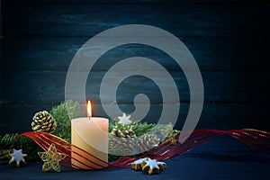 Christmas decoration with a lit white candle, fir branches, cinnamon stars and red ribbon against a dark blue wooden background,