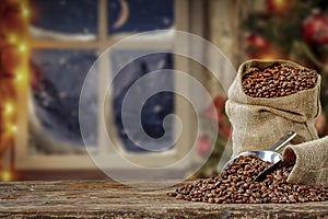 Christmas decorations and fresh coffee beans in jute sack with blurred christmas tree and sowy night window background.