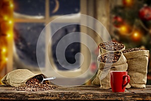 Christmas decorations and fresh coffee beans in jute sack with blurred christmas tree and sowy night window background.