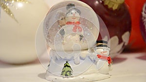 Christmas decorations. Snow falling in shiny magical crystal ball with snowman and Christmas balls in backdrop. Snowing dome