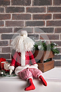 Christmas decorations, a toy Santa with a white beard sits against a brown brick wall.