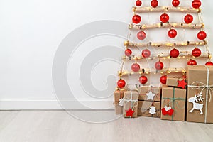 Christmas eco tree made of wooden branches hanging on white wall with festive lights and gift boxes on the floor.