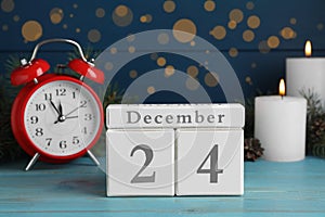 Christmas Eve - December 24. Block calendar, alarm clock and burning candles on light blue wooden table