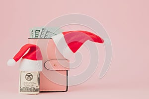 Christmas expenses. Pink leather purse with santa claus cap, gift, fir tree and dollars banknotes on pink background. Christmas