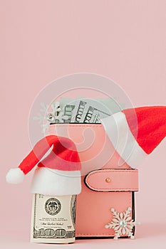 Christmas expenses. Pink leather purse with santa claus cap, gift, fir tree and dollars banknotes on pink background. Christmas