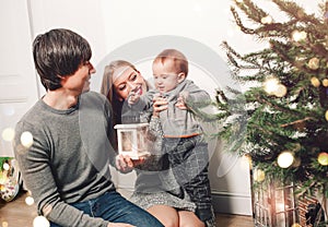 Christmas family with baby smiling near the Xmas tree. Living room decorated by Christmas tree and present gift boxes, the light