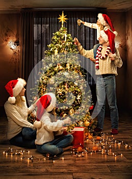 Christmas Family Decorate Xmas tree with Toys. Parents and Kids in Santa Hats together around Fir tree in cozy Living Room