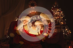 Christmas Family opening Santa Bag with Gifts. Happy Parents and Kids sitting in front of Decorated Xmas Fir Tree and Fireplace