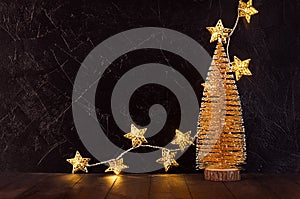 Christmas festive interior with gold Christmas tree, glowing lights with stars on black plaster wall and brown table, copy space.