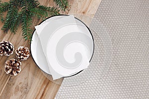 Christmas festive still life. Blank paper sheet, greeting card mockup on white plate. Old wooden bench with pine cones