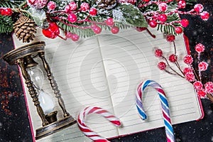 Christmas fir tree branches, decorations, candy canes, frozen red berries, cone and vintage hourglass frame on notebook with snow,