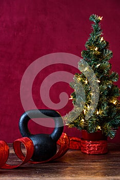 Christmas fitness, black kettle bell, with artificial Christmas tree and white lights, red ribbon, wood table, red background