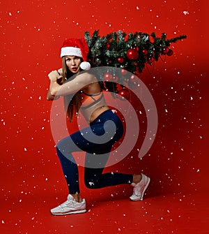 Christmas fitness sport woman wearing santa hat holding xmas tree on her shoulders. snowflakes