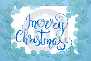 Christmas flat lay: lettering text `Merry Christmas` andblue confetti on blue background.