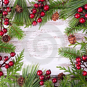 Christmas frame with evergreen branches, red berries and pine cones on a rustic square white wood background