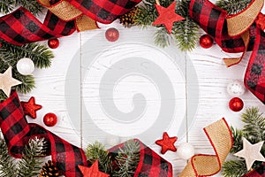 Christmas frame of ornaments, branches, burlap and red and black buffalo plaid ribbon, overhead view on a white wood background