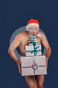 Christmas Freestyle. Young Santa Claus naked muscular body in hat standing isolated on blue covering groin with present