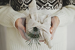 Christmas Furoshiki gift. Christmas gift wrapped in fabric in hands of woman in cozy sweater in white scandinavian room.