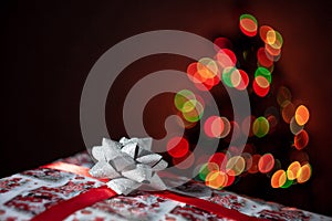 Christmas gift box or present with red bow ribbon on christmas tree bokeh background with multicolored lights. Shallow