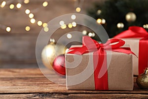 Christmas gift box with red ribbon and golden bauble on wooden table against blurred lights, space for text