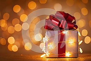 Christmas gift box with red ribbon and lights against golden bokeh background. Holiday greeting card