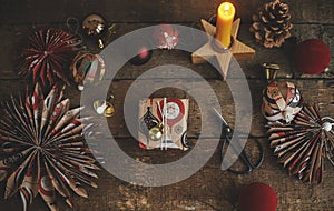 Christmas gift rustic flat lay. Stylish wrapped gift box, scissors, candle, red paper stars, golden ornaments on rustic wooden
