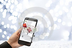 Christmas gifts in shopping cart,online purchase,hand with phone on winter snow background