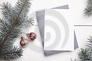 Christmas greeting card mockup with envelope, green fir tree branches and balls on white wooden background, top view, flat lay.