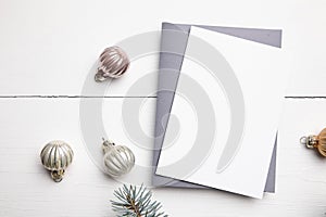 Christmas greeting card mockup with envelope, green fir tree branches and balls on white wooden background, top view, flat lay.