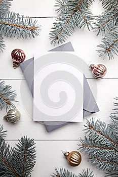 Christmas greeting card mockup with envelope, green fir tree branches and balls on white wooden background, top view, flat lay.