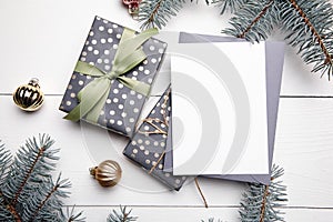 Christmas greeting card mockup with grey envelope, gift boxes and decorations on white wooden background, top view, flat lay.