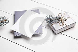 Christmas greeting card mockup with grey envelope, silver color gift box and green fir tree branch on white wooden table. Empty