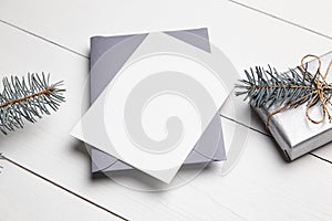 Christmas greeting card mockup with grey envelope, silver color gift box and green fir tree branch on white wooden table. Empty