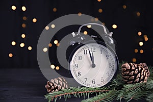 Christmas greeting card with space for text. Alarm clock, fir tree branches and pine cones on wooden table against blurred festive