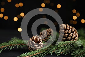 Christmas greeting card with space for text. Fir tree branches and pine cones on wooden table against blurred festive lights