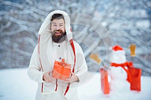 Christmas greeting card. Winter portrait of handsome hipster in snow Garden with gift make snowman. Man with red gift