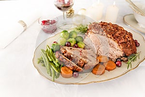 Christmas ham or roast pork with crust, green beans, Brussel sprouts, carrots and cranberry on a white table cloth with candles