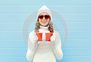 Christmas happy smiling young woman with gift box wearing knitted hat sweater sunglasses over blue background