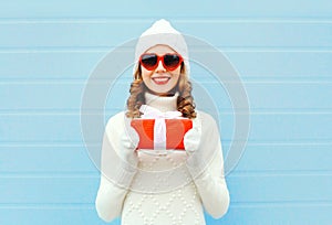 Christmas happy smiling young woman with gift box wearing a knitted hat sweater sunglasses over blue