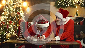 Christmas helper and Santa. Grandfather Santa and grandchild son writing wish list letter to Santa Claus.