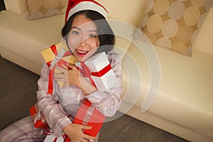 Christmas holiday lifestyle portrait at home of young beautiful and happy Asian American woman in pajamas and Santa hat  holding