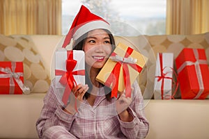 Christmas holiday lifestyle portrait at home of young beautiful and happy Asian American woman in pajamas and Santa hat  holding