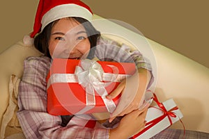 Christmas holiday portrait at home of young beautiful and happy Asian Chinese woman in pajamas and Santa hat  holding present and