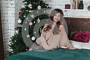 Christmas Holidays. Cute brunette girl sits on a bed. Loft bedroom decorated with Christmas garlands and fir.