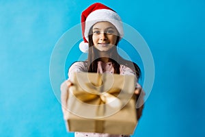 Smiling teenage girl in santa helper hat holding gift box over lights on blue background