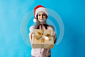 Smiling teenage girl in santa helper hat holding gift box over lights on blue background