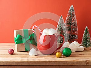 Christmas hot chocolate cup with marshmallow, gift box and decorations on wooden table over red background