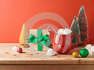 Christmas hot chocolate cup with marshmallow, gift box and decorations on wooden table over red background