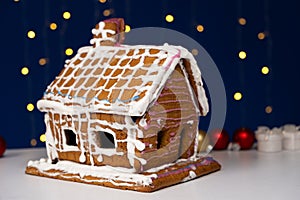 Christmas house made of gingerbread cookies decorated with sugar icing on blue background with bokeh garland